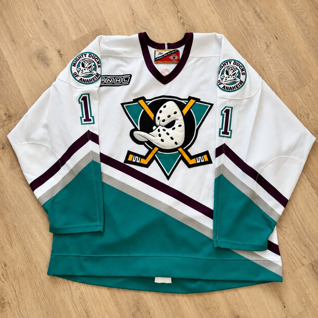Mighty Ducks of Anaheim third jersey in white with eggplant and jade accents, Anaheim shoulder patches, and #1, laid out on a wood floor.