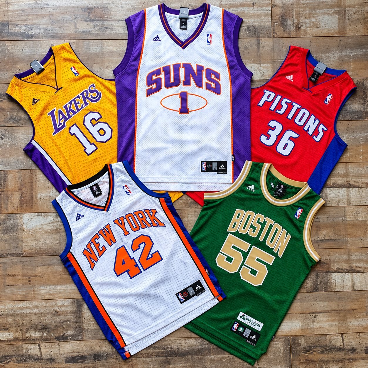 Five retro basketball jerseys laid on a wooden floor including Lakers #16, Suns #1, Pistons #36, Knicks #42 and Celtics #55